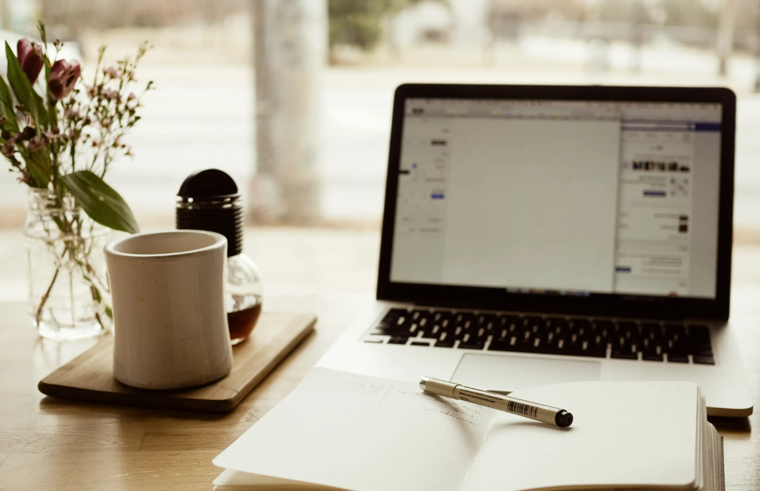 Laptop and open notebook with pen on a wooden desk next to a coffee mug and flowers, showing a calm setup for writing evergreen content
