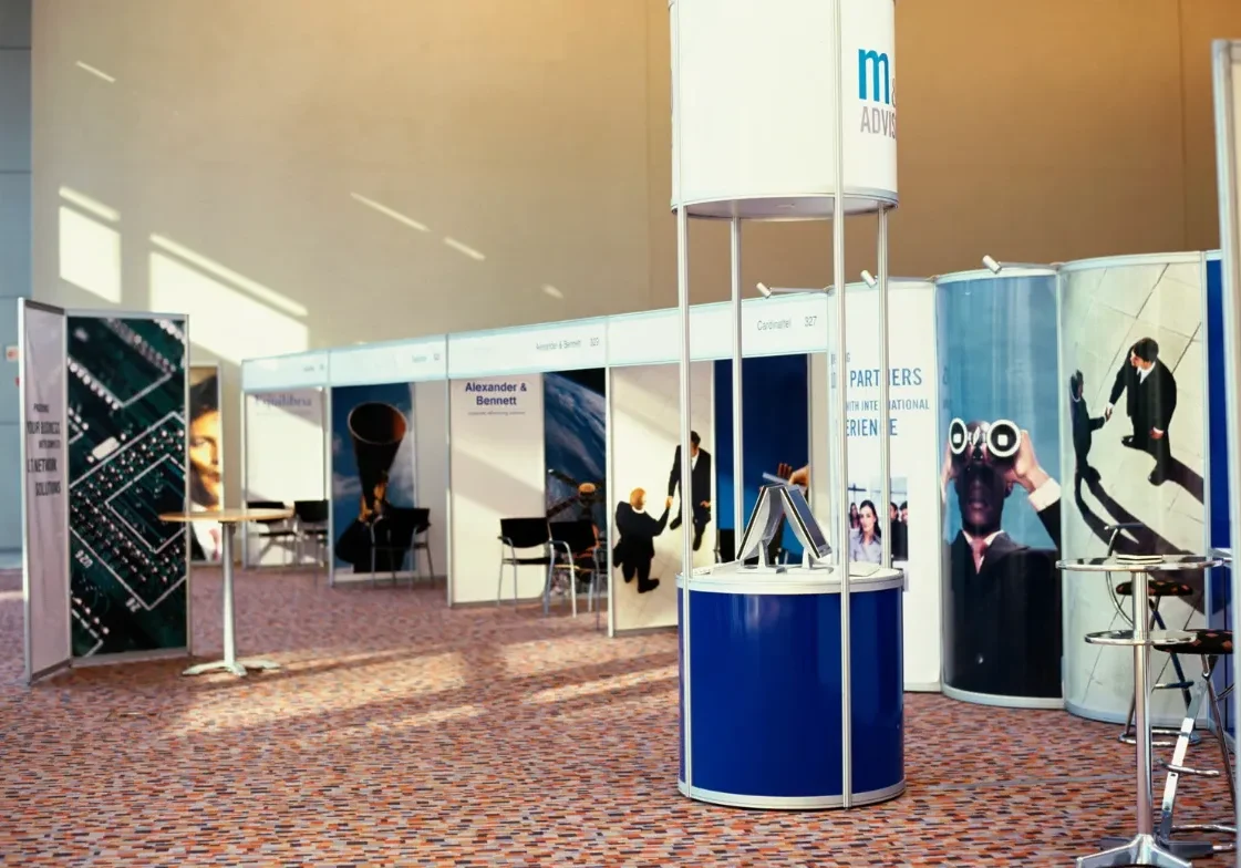 Indoor exhibition hall featuring modular booths, a central circular display stand, branded graphics and high tables under natural light